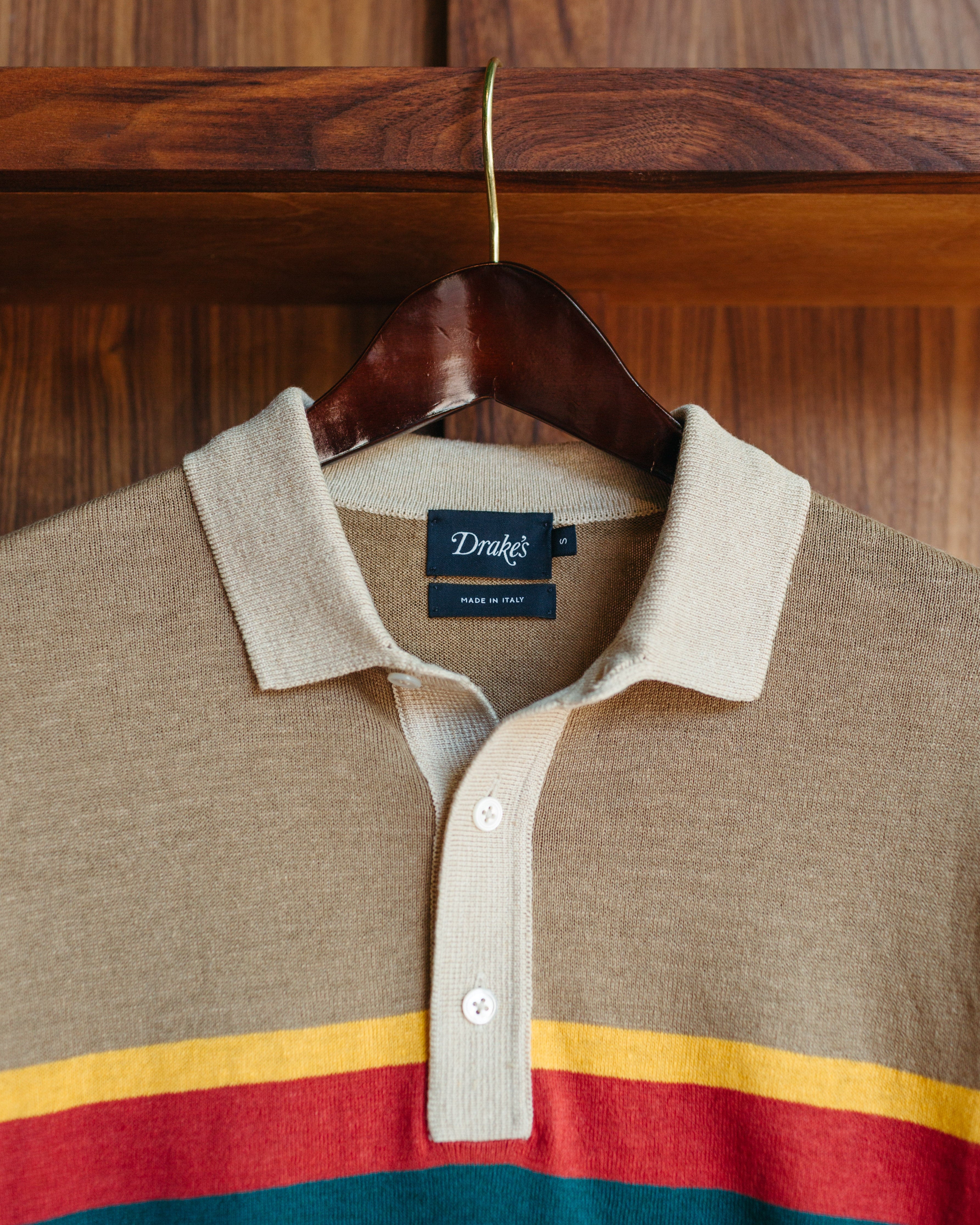 A beige knit polo shirt with horizontal stripes in yellow, red, and teal, hanging on a dark wooden hanger against a wooden background. The brand label reads 'Drake's'.