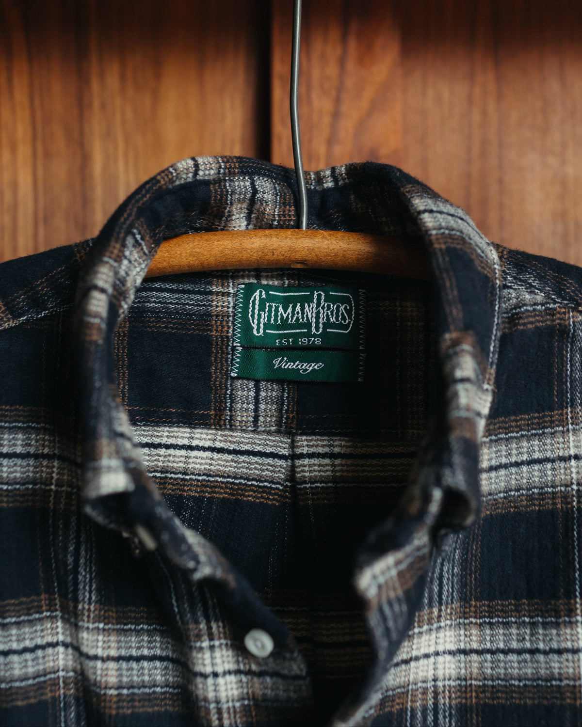 A vintage plaid tweed flannel shirt by Gitman Vintage featuring a dark navy and brown plaid pattern, hanging on a wooden hanger against a wooden background.