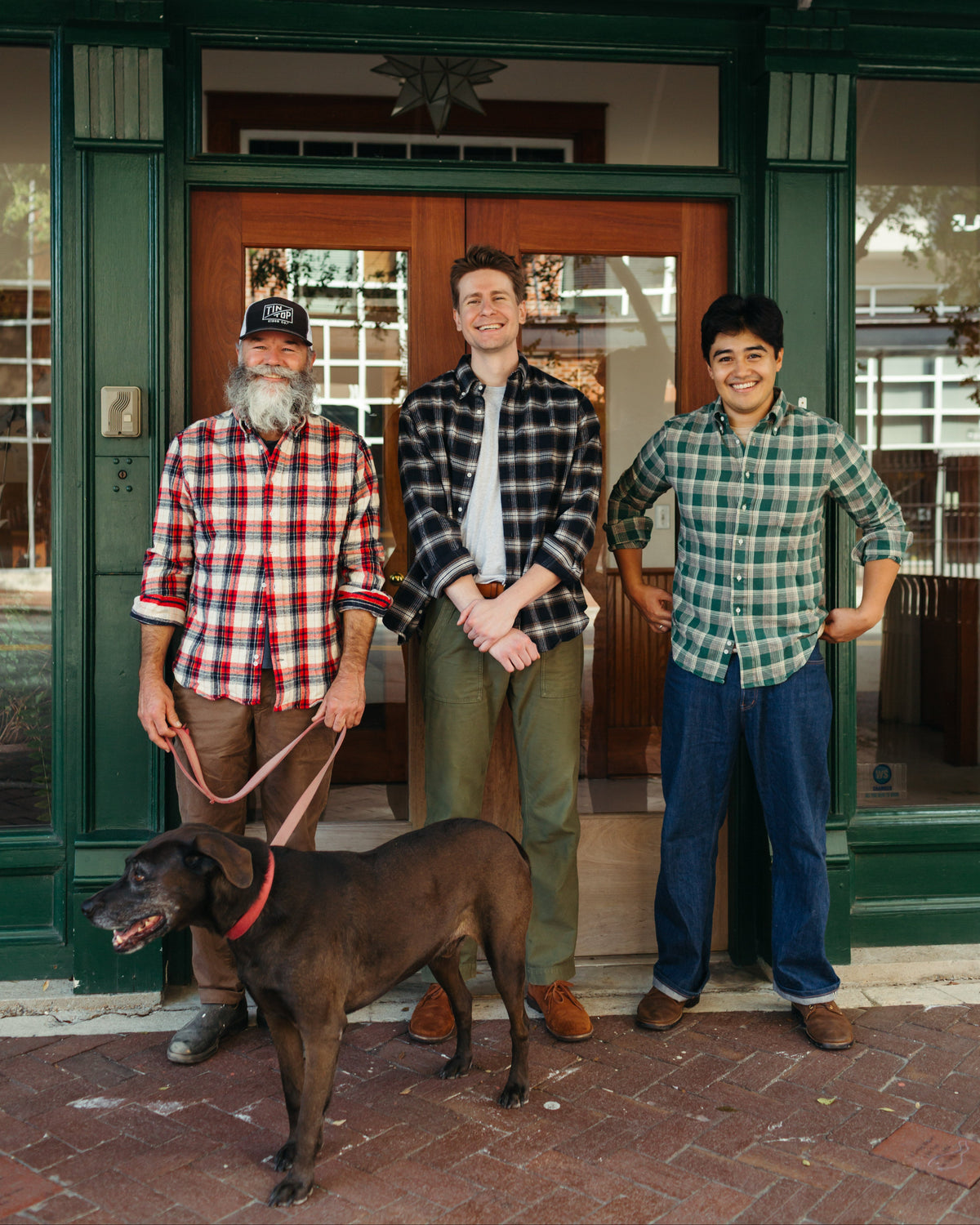 Three men standing in front of a green storefront doorway with a large brown dog on a leash in the foreground. The men are wearing long-sleeve plaid Gitman Vintage flannel shirts in red, black-and-white, and green, paired with jeans and pants.