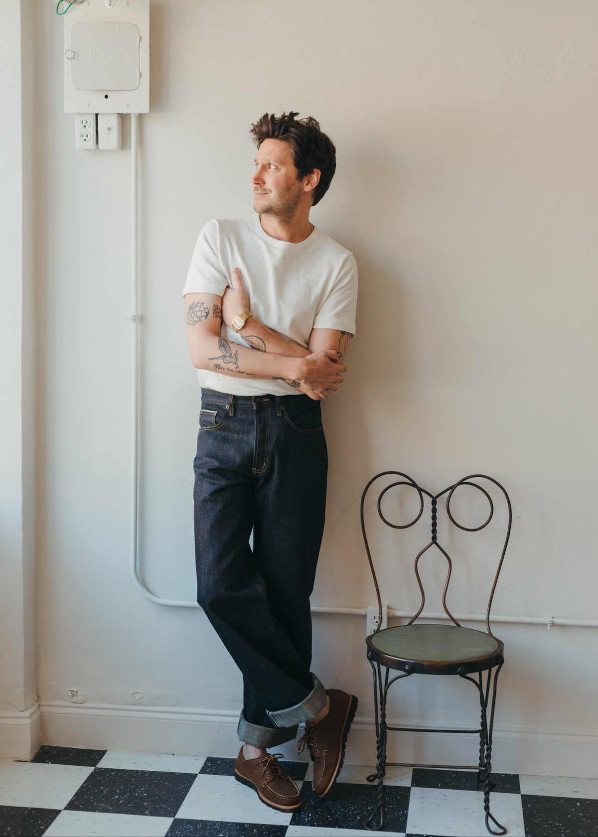 A man with tattoos leans against a cream-colored wall in a room with a black and white checkered floor. He is wearing a white Merz B Schwanen heavyweight tee, Naked & Famous Harionago Selvedge denim in the strong guy fit, and brown leather loafers from Rancourt & Co.