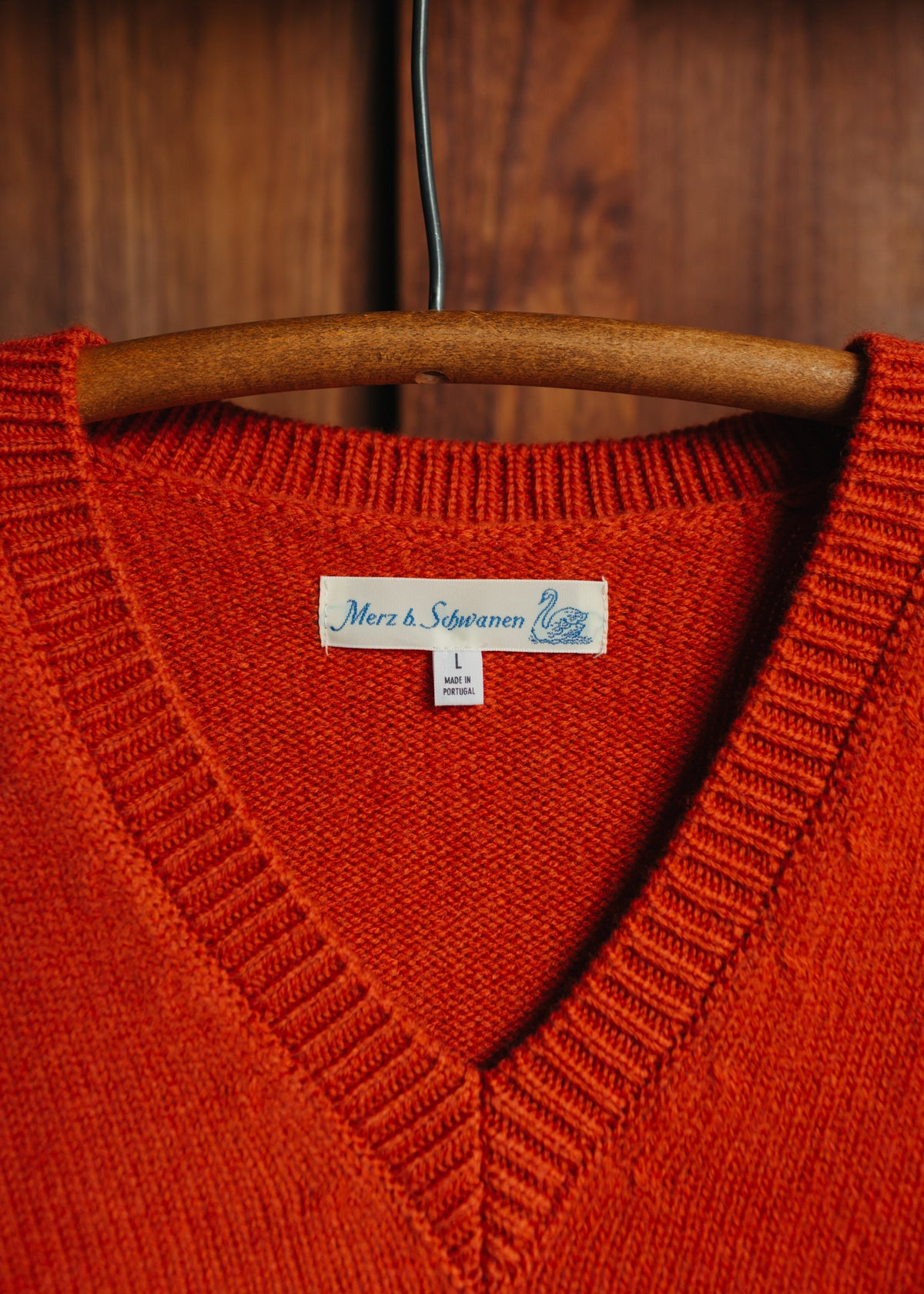 A close-up of a vibrant orange-red knit sweater from Merz B Schwanen hanging on a wooden hanger against a dark wood background. The brand label 'Merz & Slevanen' is visible inside the V-neck.