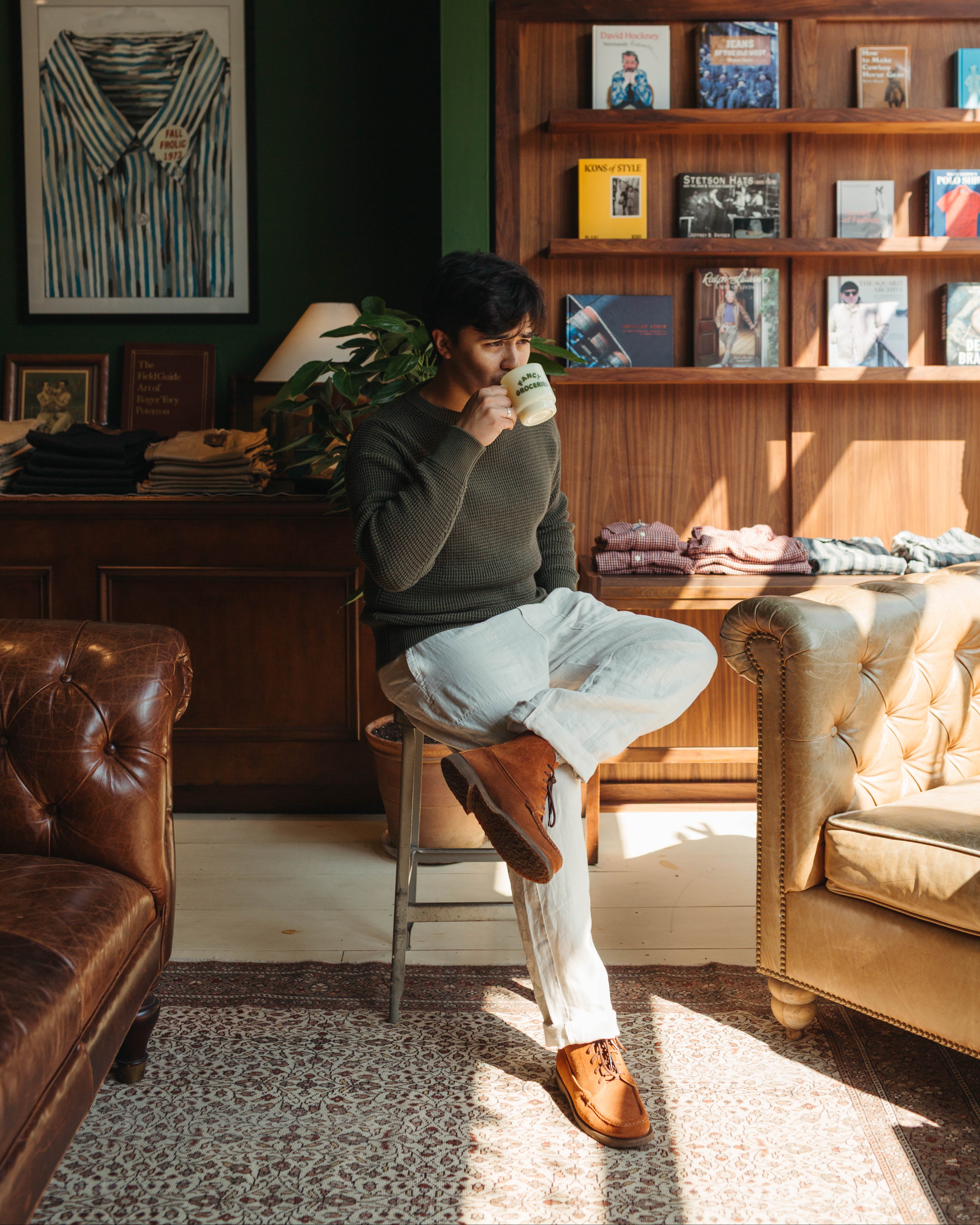 A man sits on a metal stool in a cozy, sunlit room, at Fancy Groceries in Winston Salem NC, wearing a dark olive green sweater, off-white trousers, and brown leather shoes while holding a mug. The background features a dark wood bookshelf, a green wall, and leather furniture.