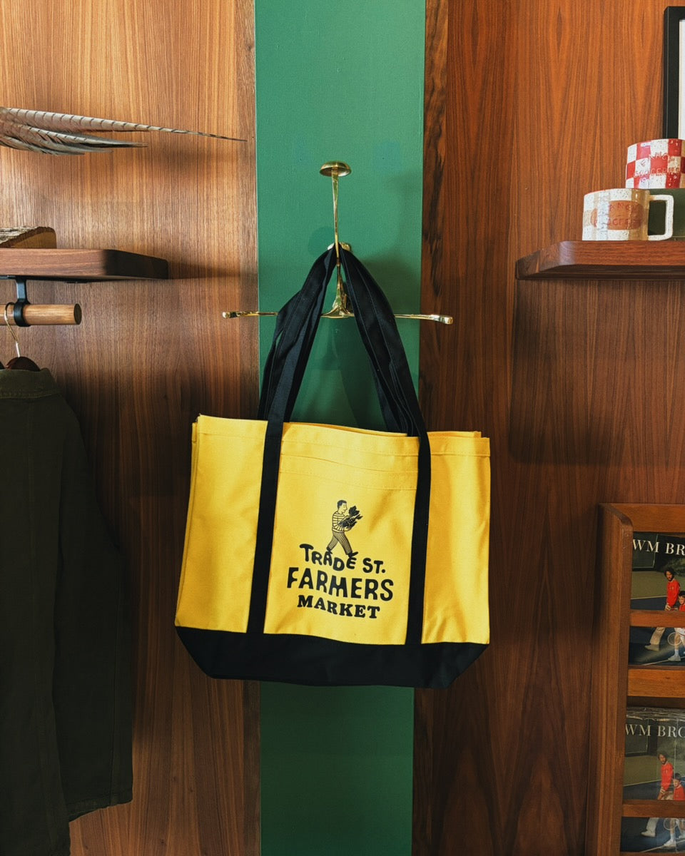 Yellow tote bag with black accents hanging on a green wall next to wooden shelves. Fancy Groceries. Trade Street Farmers Market Tote Bag.