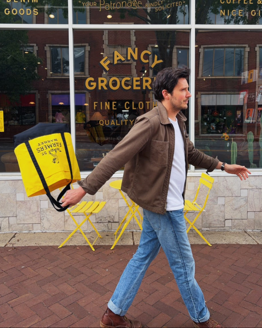 Man walking past a store with a yellow bag, in front of a 'Fancy Grocer' sign.