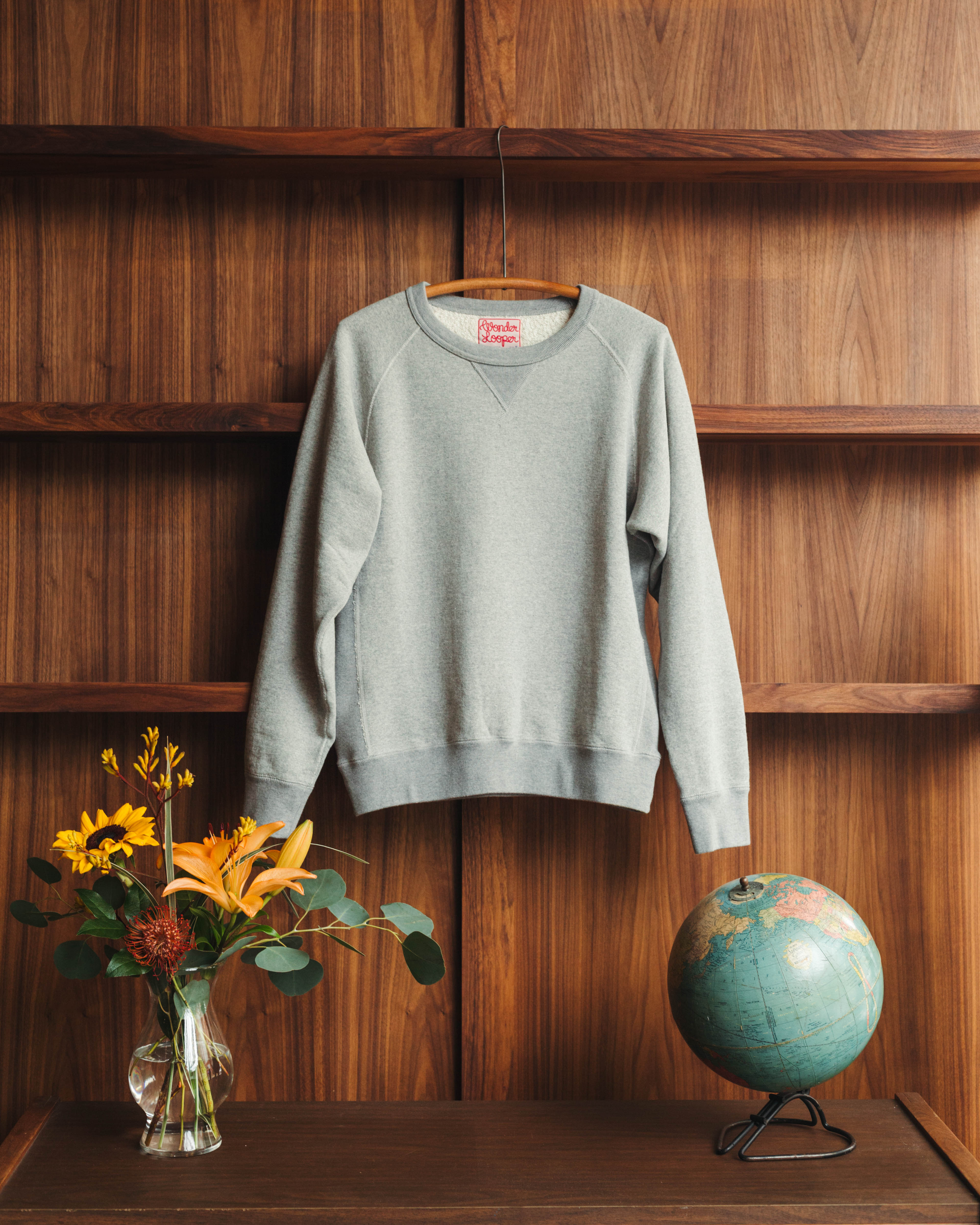 Gray sweatshirt on a wooden hanger against a wooden wall with a vase of flowers and a globe. Wonder Looper at Fancy Groceries in Winston Salem.