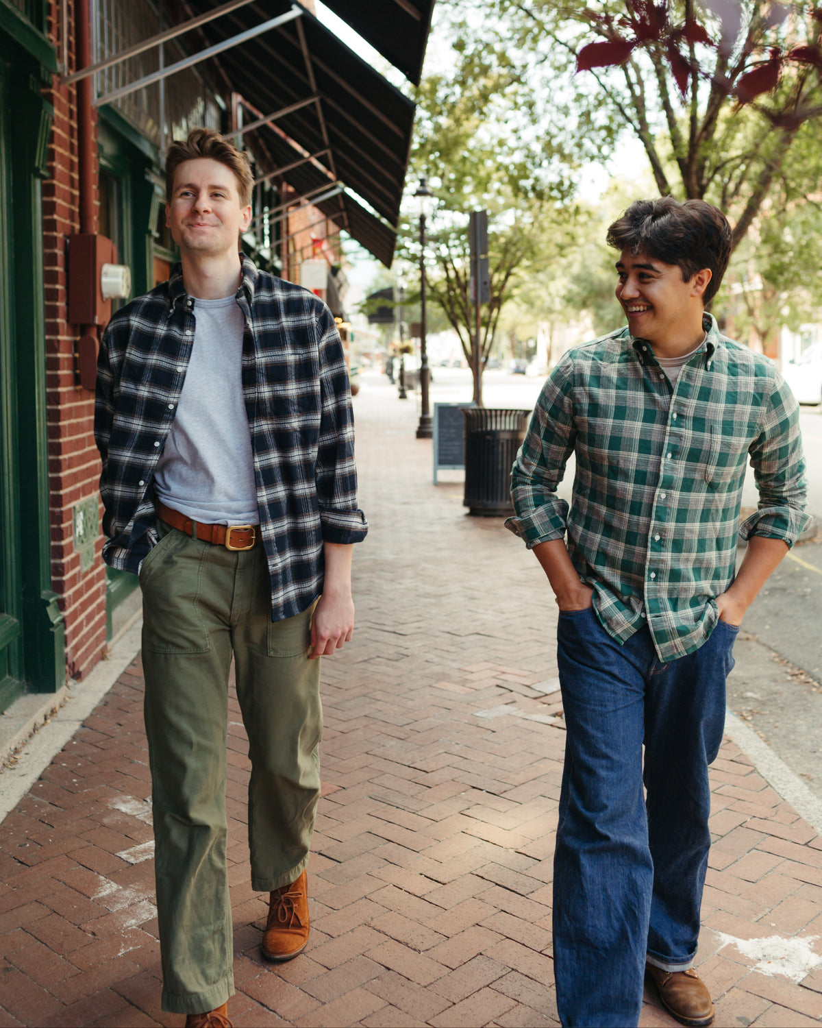 Two men walking side-by-side on a brick sidewalk in an outdoor urban setting. The man on the left wears a black and white plaid flannel shirt over a white t-shirt, olive green pants, and brown boots.