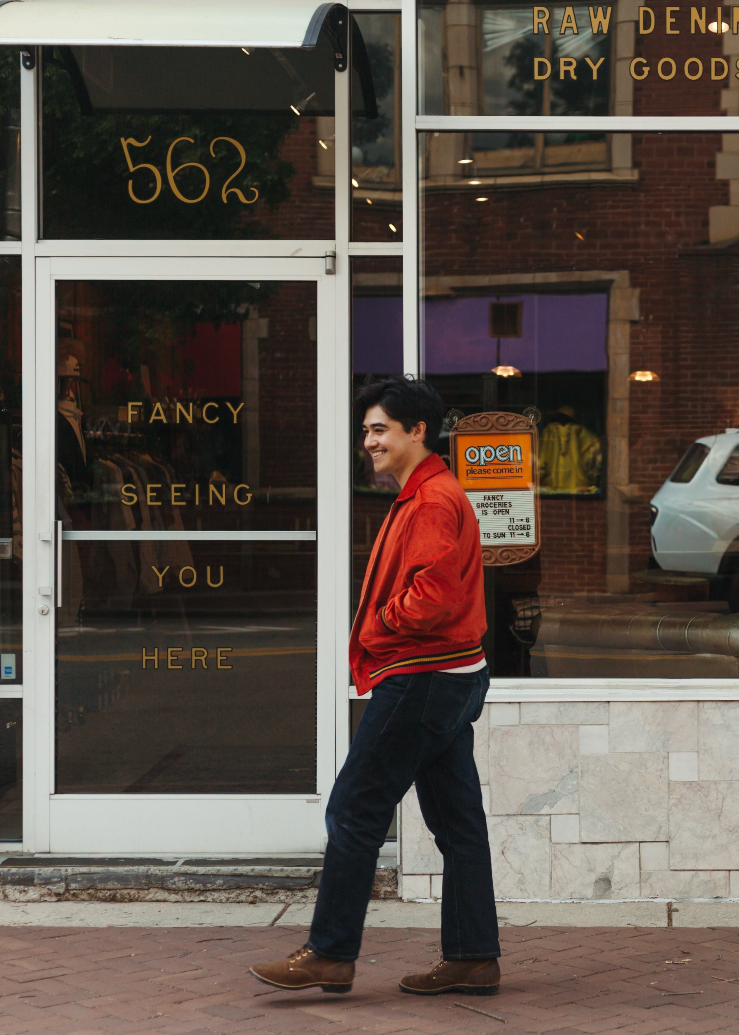 Man in a red jacket walking past a store with a sign for 'Fancy Groceries' and 'Fancy Seeing You Here' wearing Dehen1920