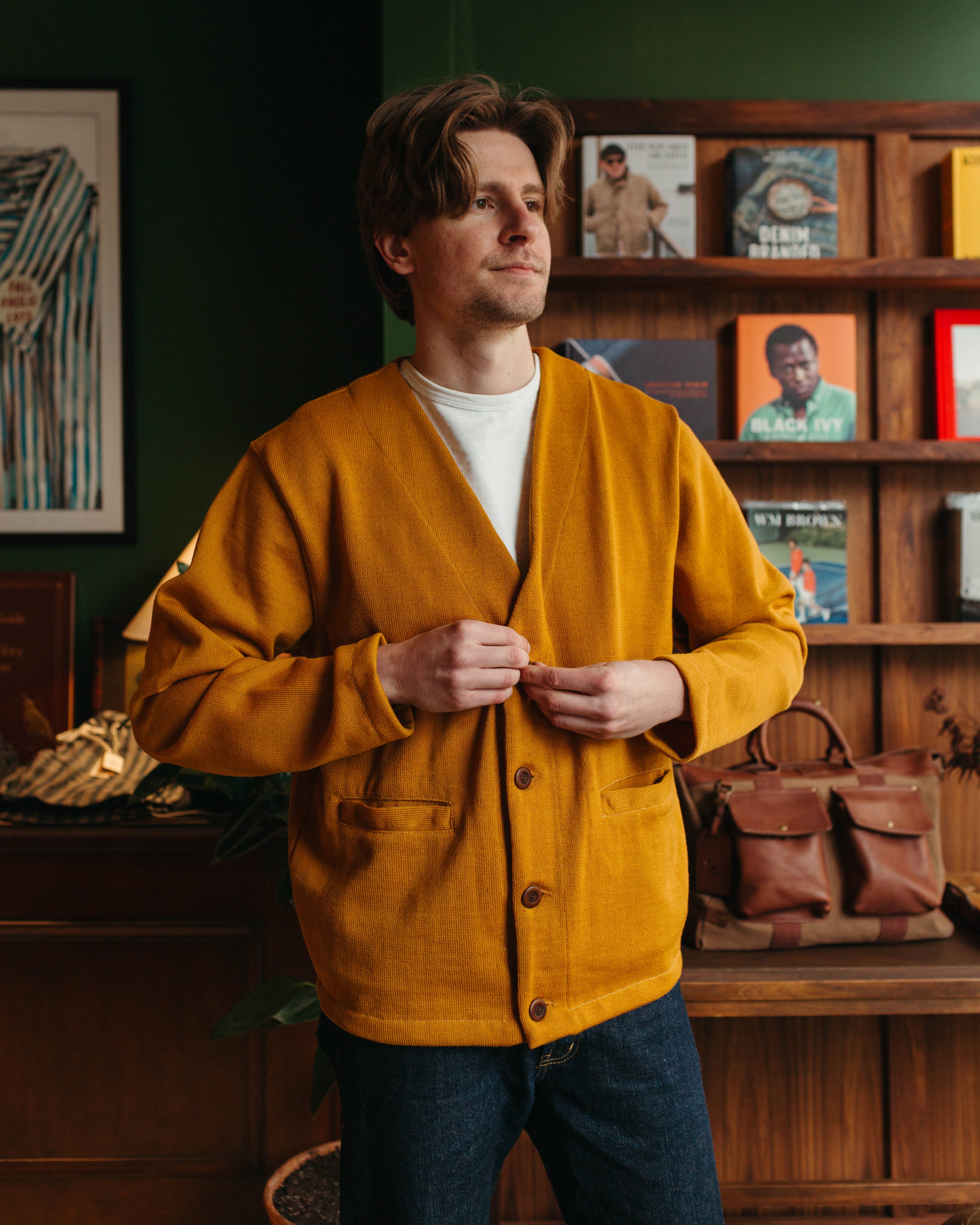 A man wearing a mustard yellow Dehen1920 wool cardigan over a white t-shirt and dark blue jeans at Fancy Groceries, standing in a warm, rustic room with wooden bookshelves in the background.