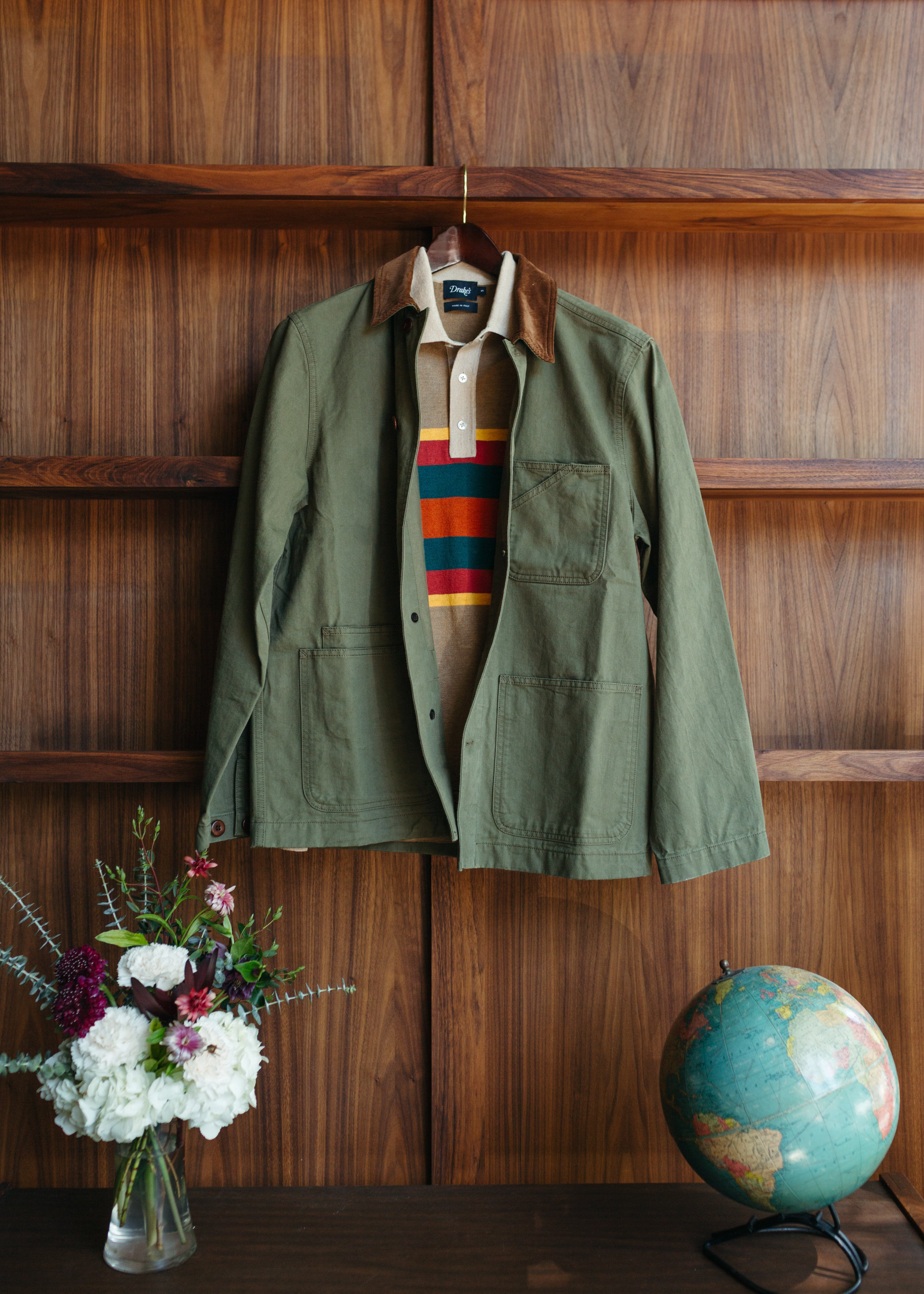 An olive green utility jacket with brown corduroy collar detail, hanging on a wooden hanger against a dark wood-paneled wall. Underneath, a beige collared shirt with red, blue, and yellow horizontal stripes is visible.