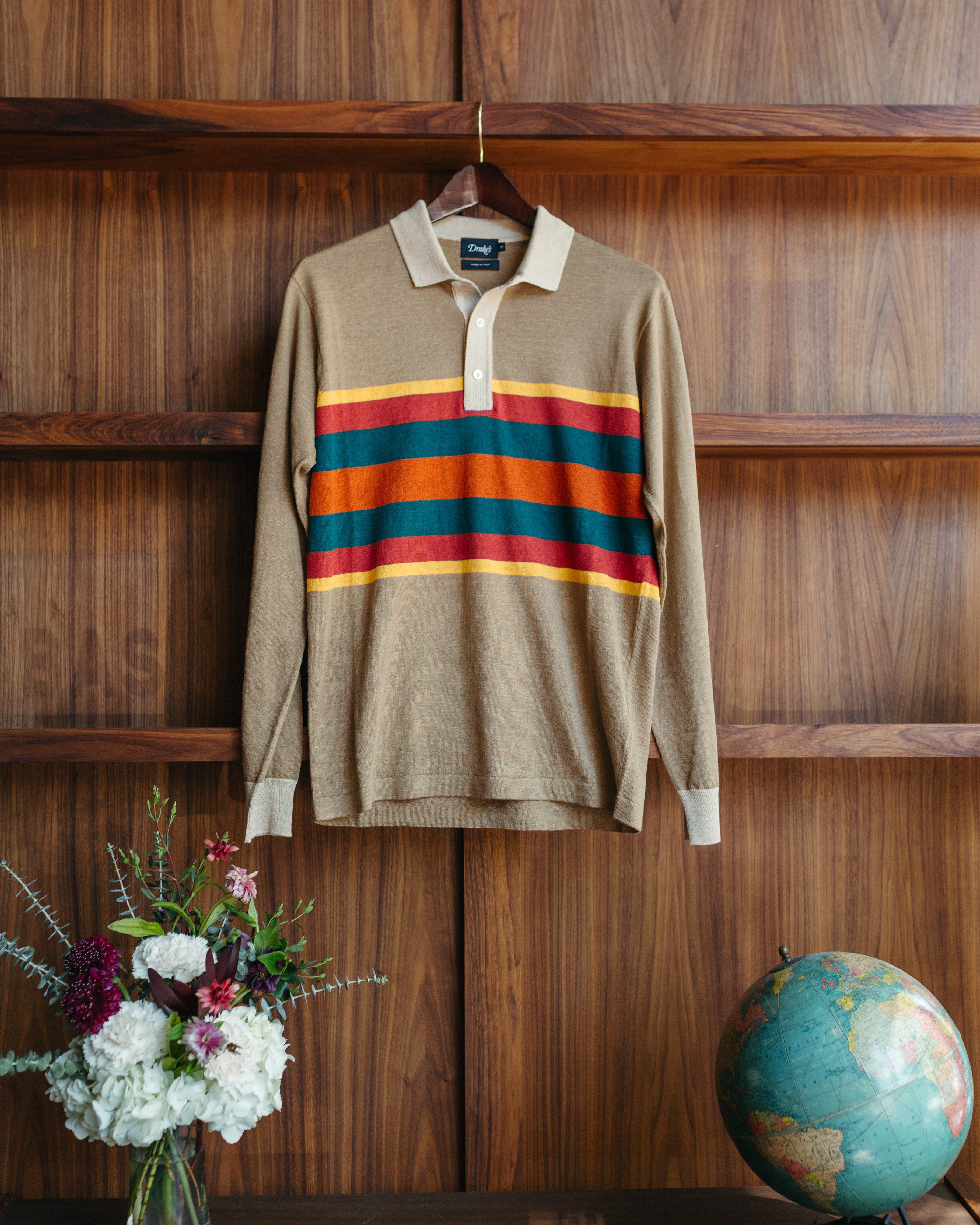 A Drake's beige long-sleeve rugy polo sweater hanging at Fancy Groceries with horizontal stripes in red, orange, and yellow across the chest, hanging on a wooden hanger against a dark wood paneled wall. A vase of flowers and a globe sit on the table below.