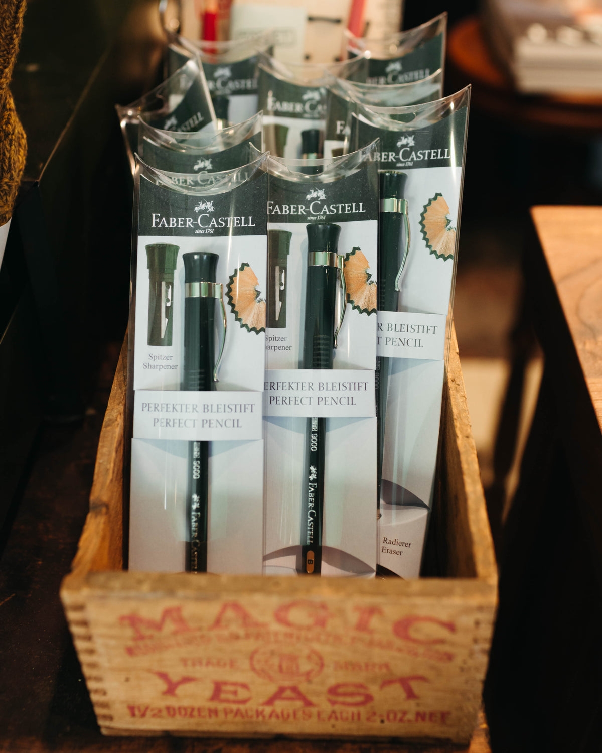 Several Faber-Castell Perfect Pencil packages arranged inside a wooden crate on a dark tabletop at Fancy Groceries