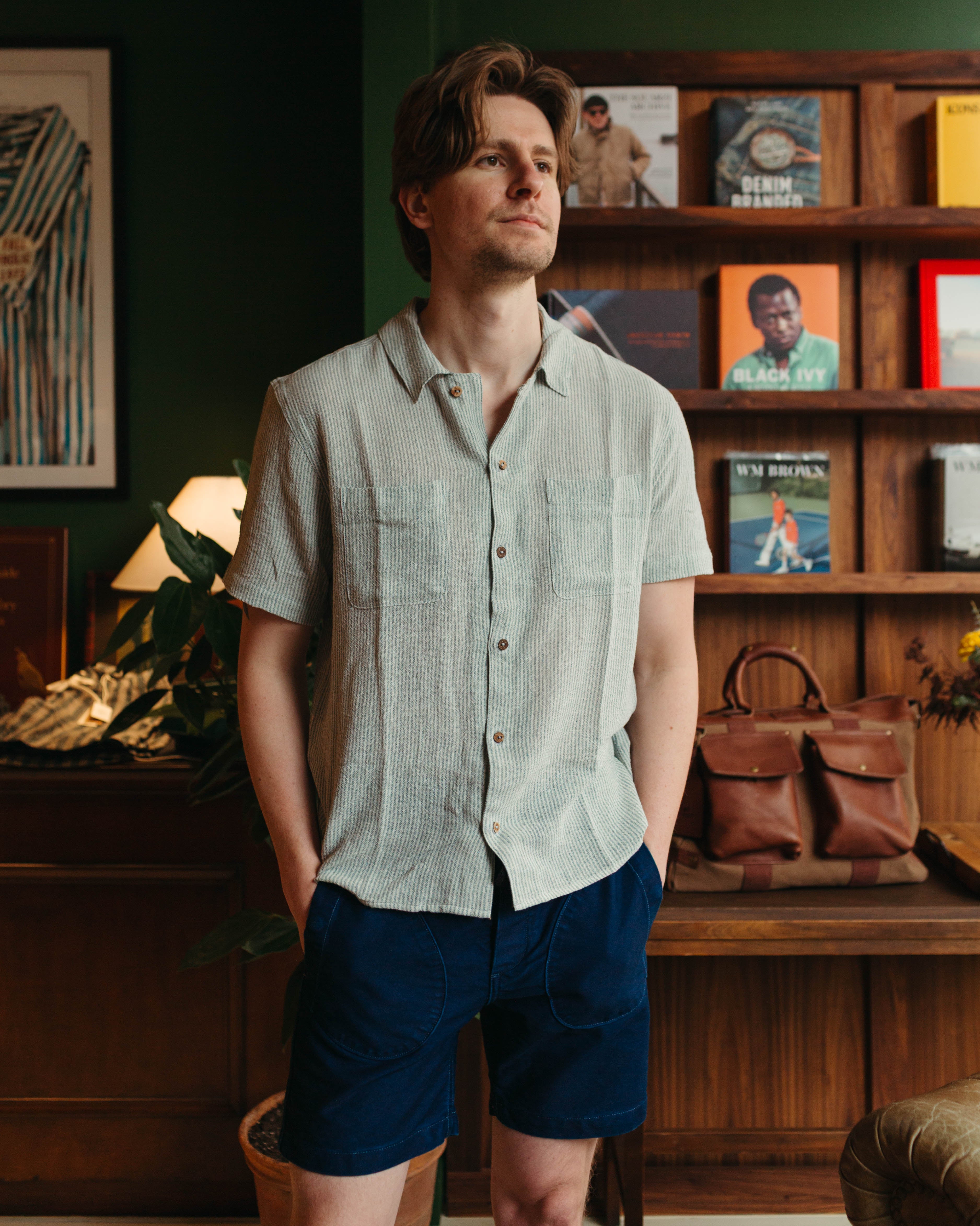 A man wearing a light sage green short-sleeve button-up shirt and navy blue shorts, standing in a warm, dark wood-paneled room with bookshelves and a leather bag in the background.