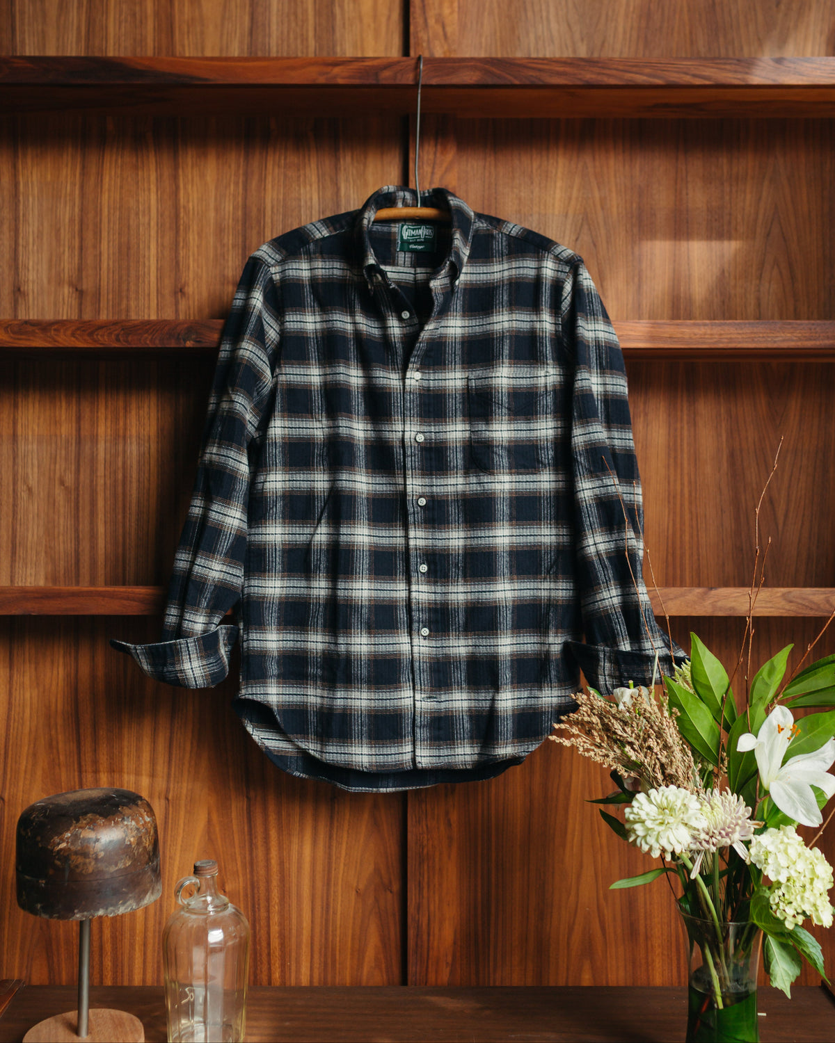A long-sleeve plaid button-up shirt by Gitman Vintage in dark navy and gray hangs on a wooden hanger against a warm wood-paneled wall at Fancy Groceries