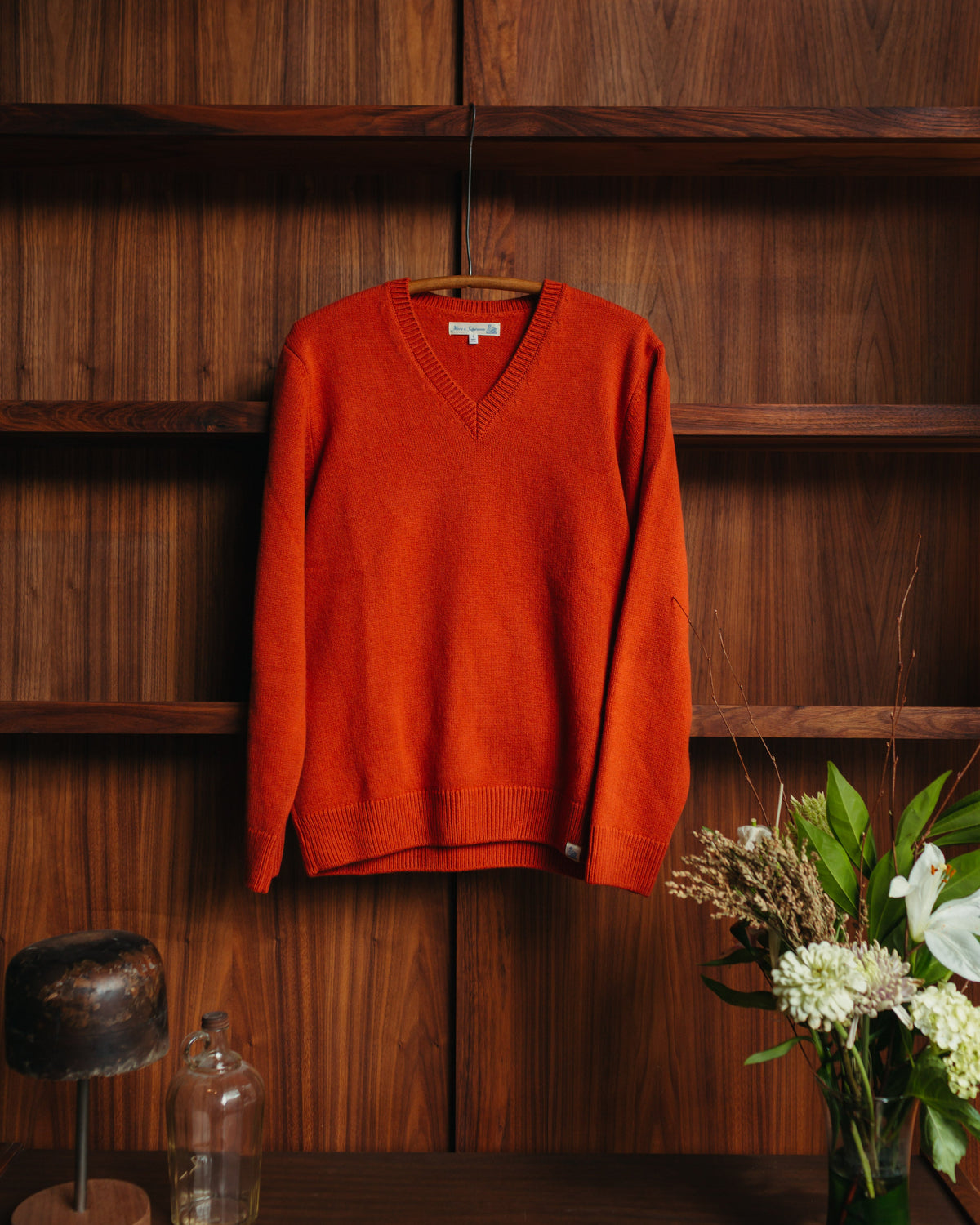 A rust-orange v-neck knit sweater from Merz B Schwanen hanging on a wooden hanger at Fancy Groceries against a dark wood-paneled wall, with a vase of white flowers and a small glass bottle on a table below.