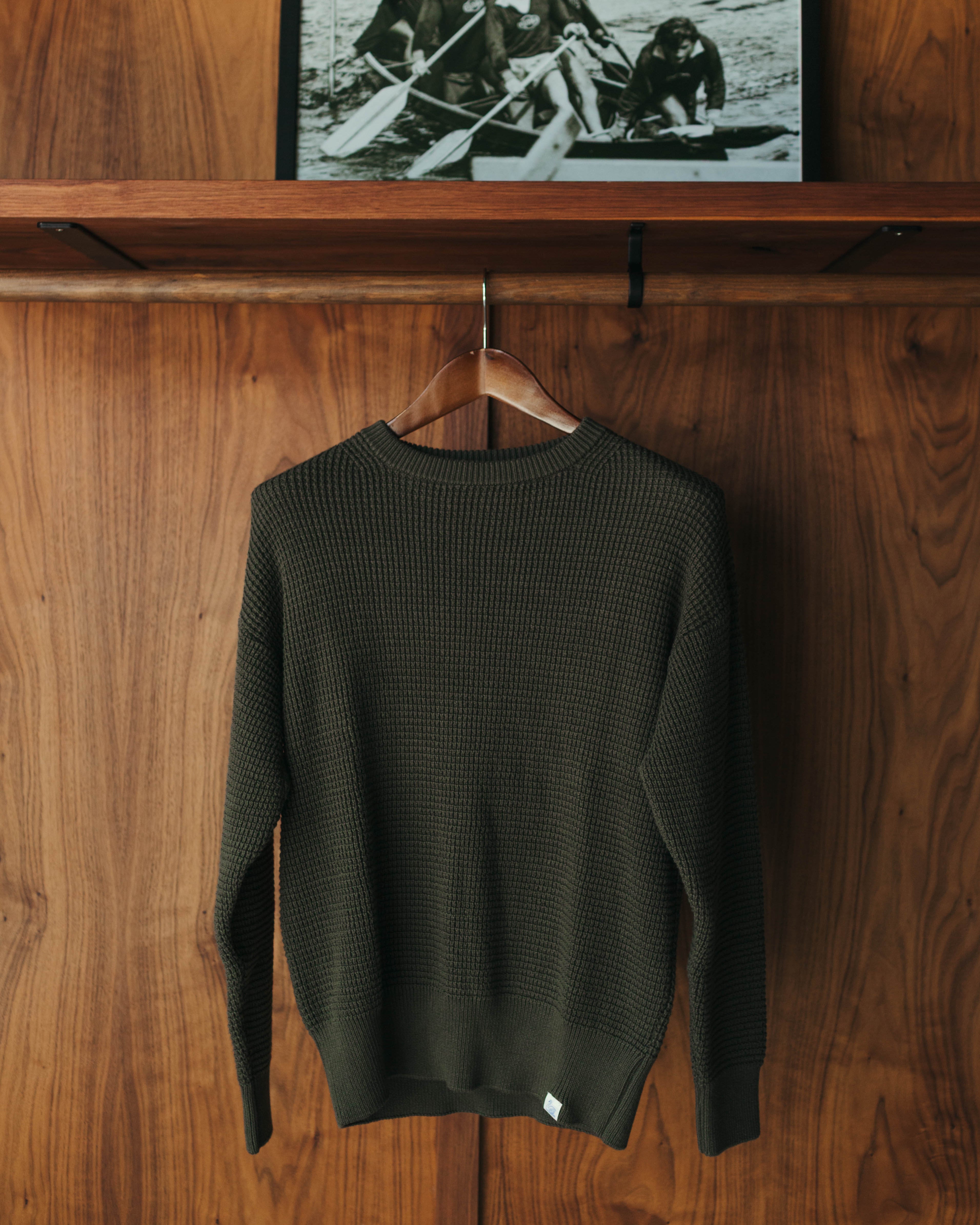 An olive green textured knit sweater from Merz B Schwanen hangs on a wooden hanger at Fancy Groceries against a warm wood-paneled wall, with a framed black-and-white photo displayed on a wooden shelf above.