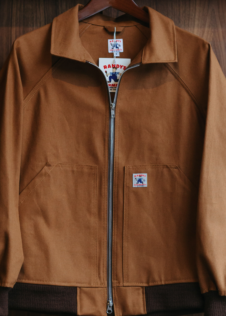 A brown suede zip-up jacket with a high collar and front pockets, hanging on a wooden hanger against a dark wood-paneled background. The jacket features a small embroidered patch on the left chest and a visible 'SAPPHIRE' brand tag at the collar.