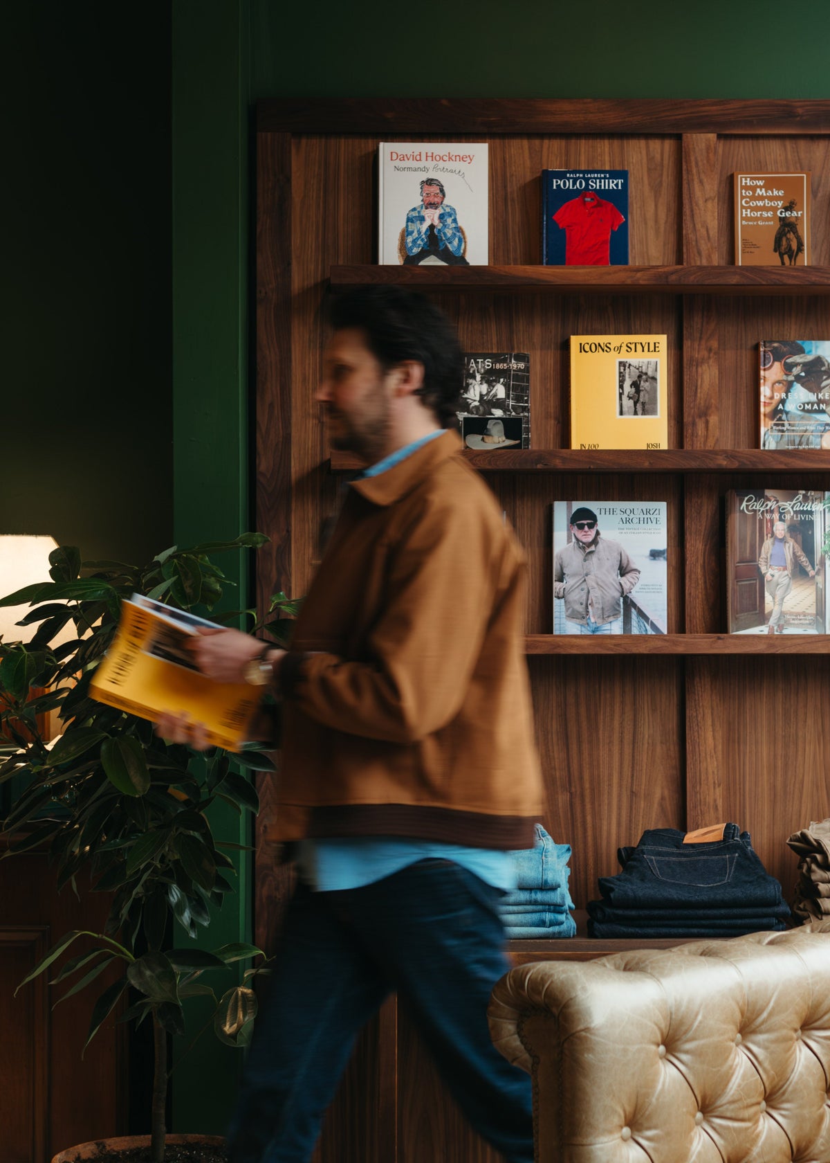 A man in a brown Randy's Garments 12oz Canvas Jacket and blue jeans walks past a wooden bookshelf at Fancy Groceries filled with books and clothing inside a stylishly decorated room with dark green walls and a tufted beige sofa.