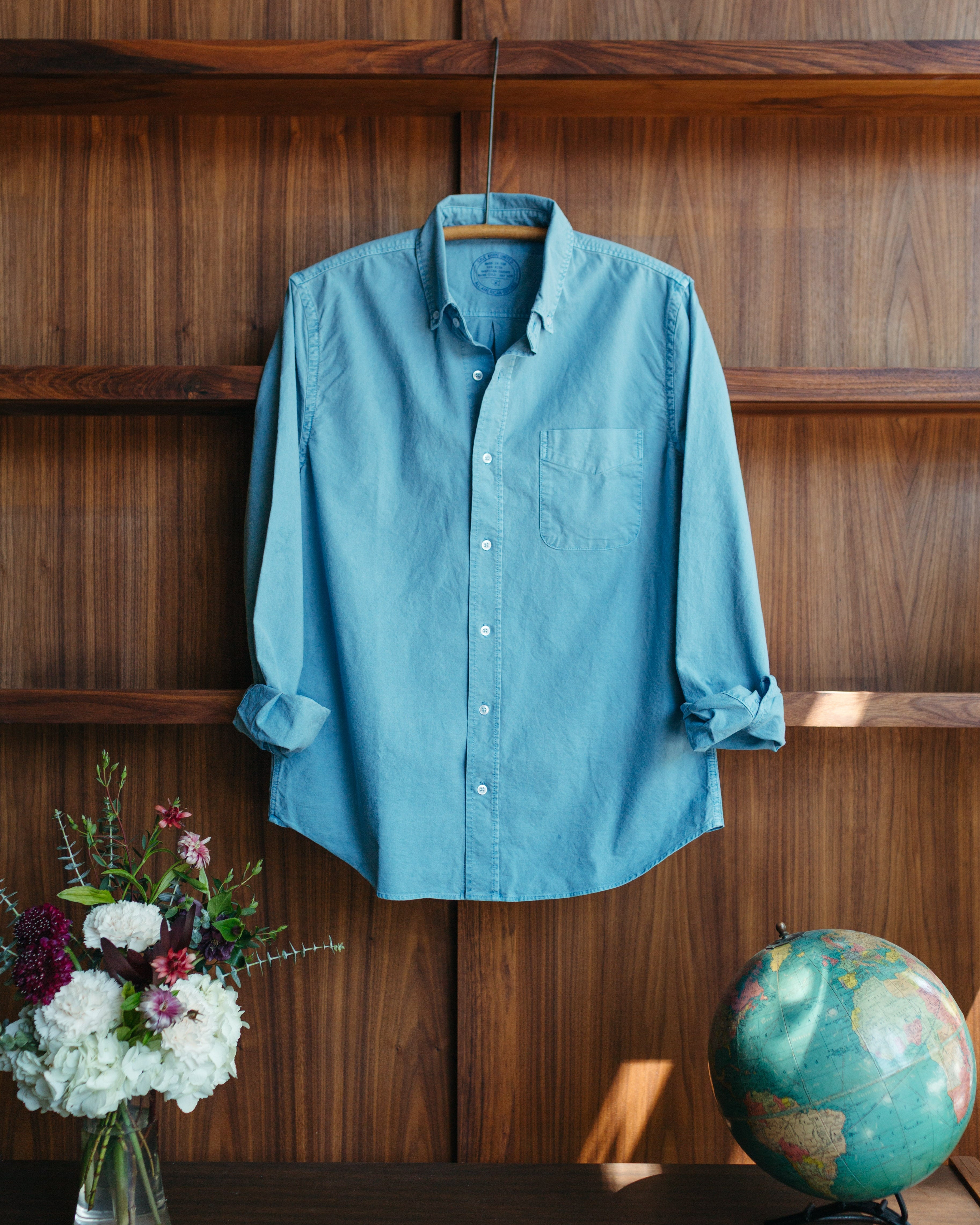 Save Khaki All American Oxford in Ocean - a light blue long-sleeve button-up shirt hanging on a wooden hanger against a dark wood-paneled wall, with a vase of flowers on the left and a globe on the right.