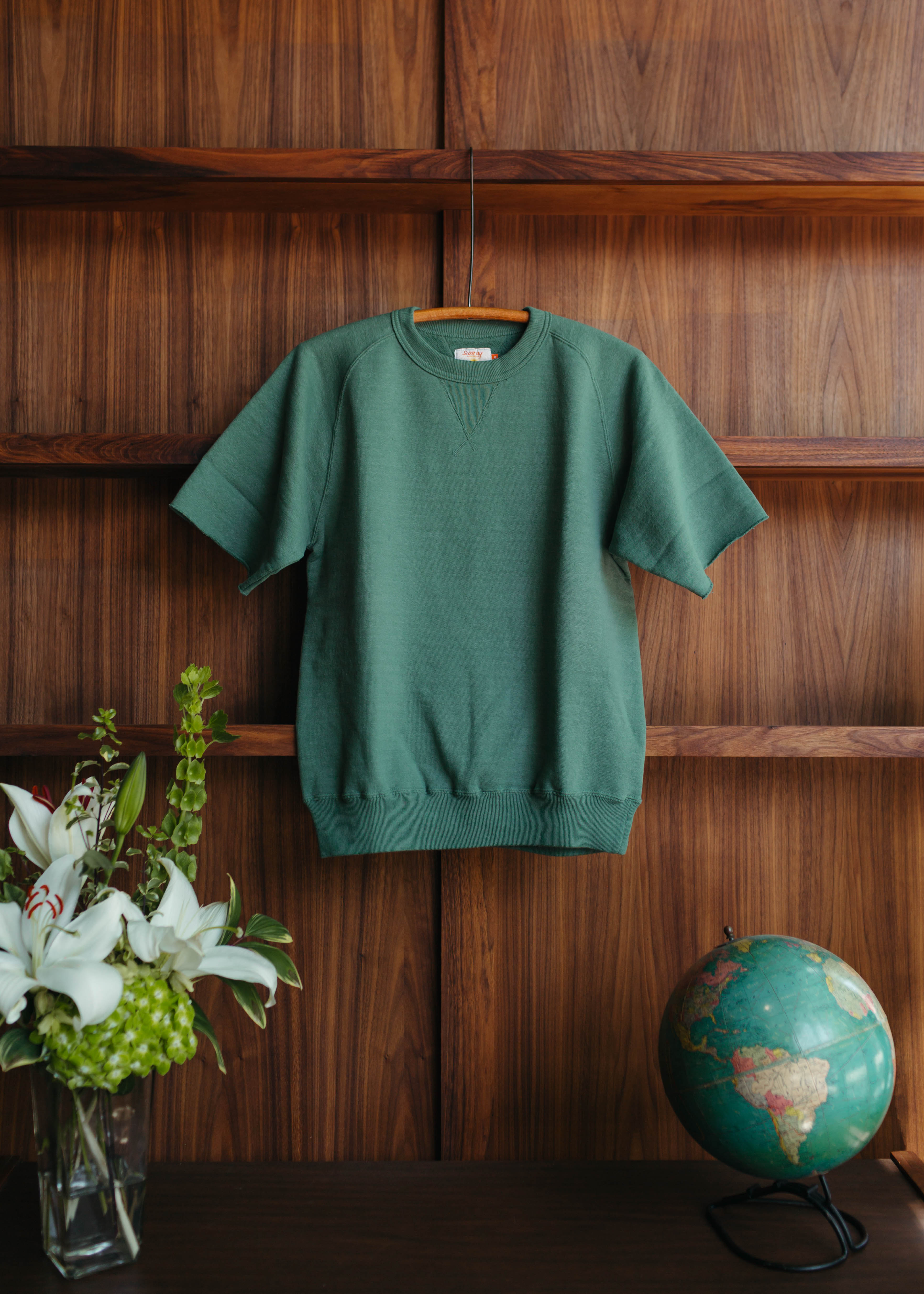 Sunray Green sweater on a wooden hanger against a wooden wall with a vase of flowers and a globe at fancy groceries.