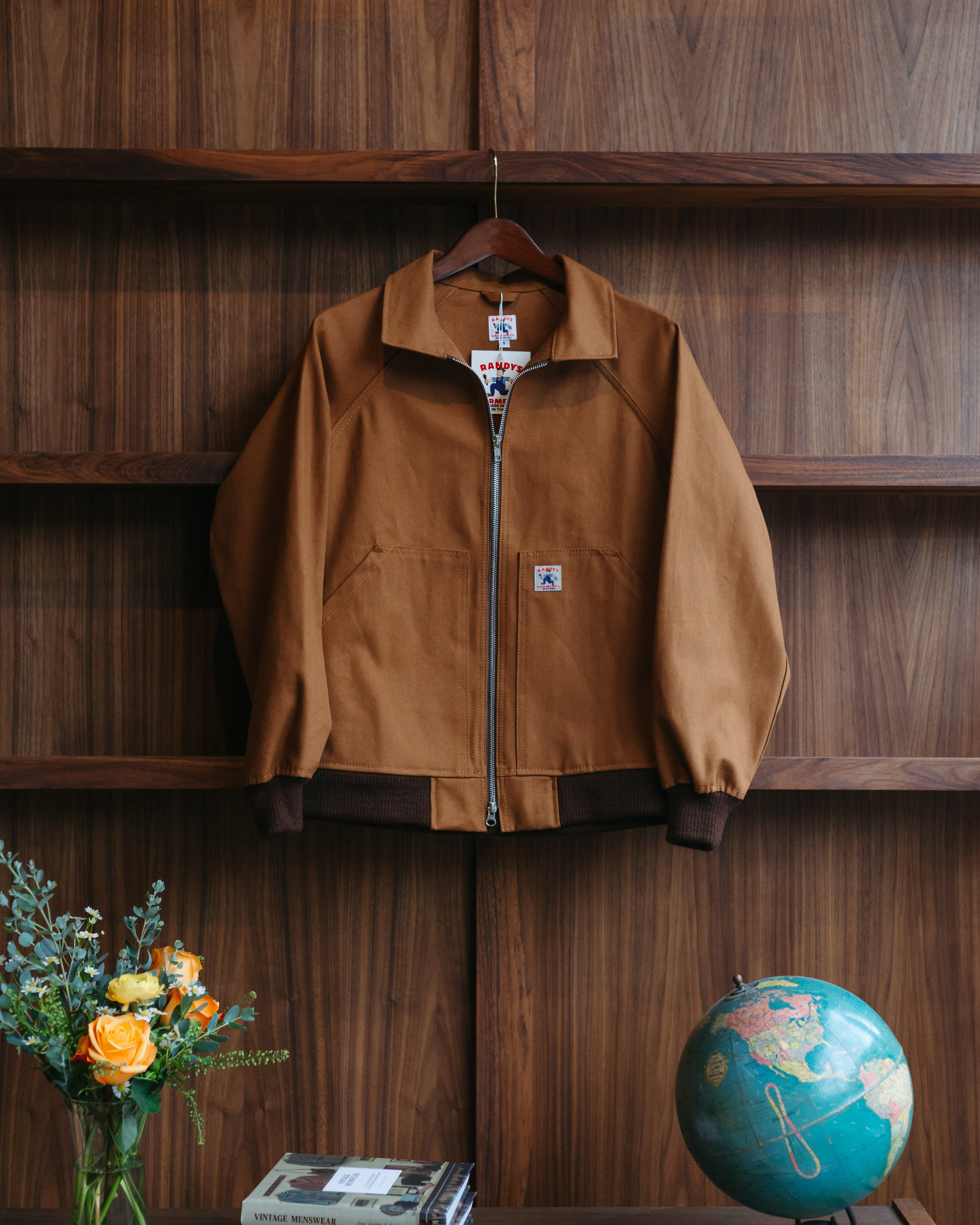 A brown Carhartt style jacket from Randy's Garments hanging at Fancy Groceries against a dark wood-paneled wall, with a vase of flowers, a stack of books, and a globe on a table below.
