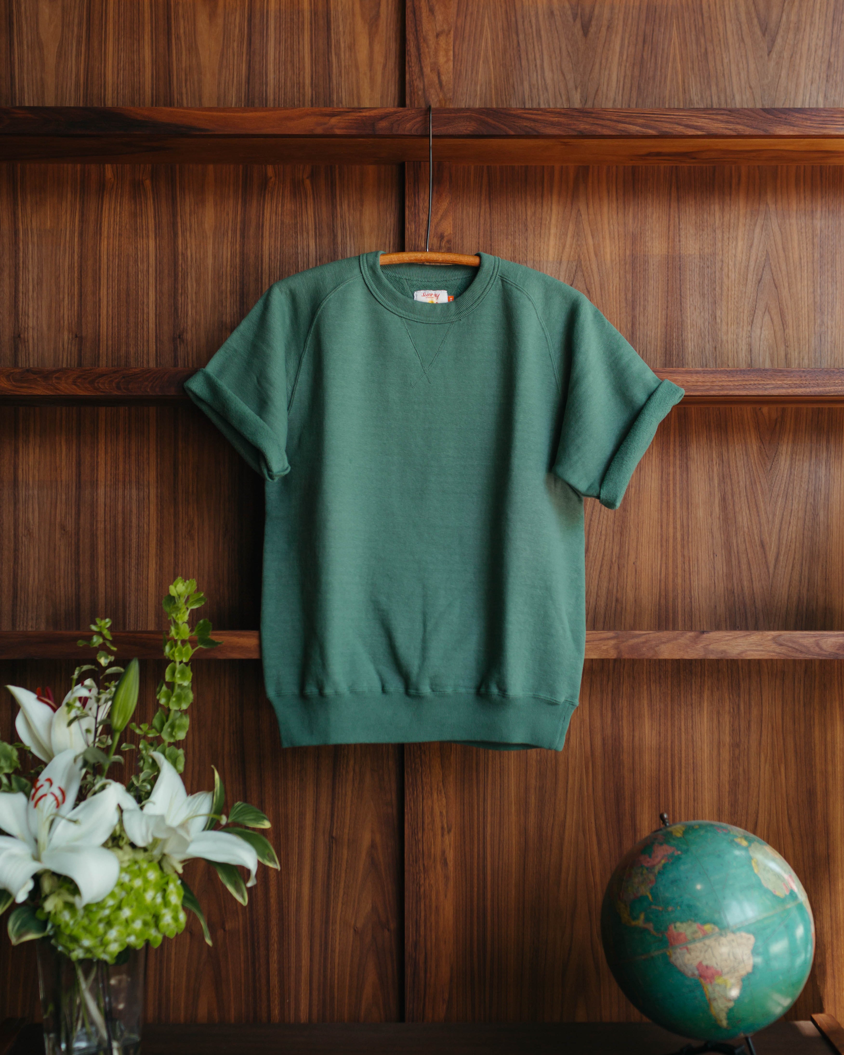 Green Sunray Sportswear short sleeve sweatshirt on a wooden hanger against a wooden wall with a vase of flowers and a globe at Fancy Groceries.