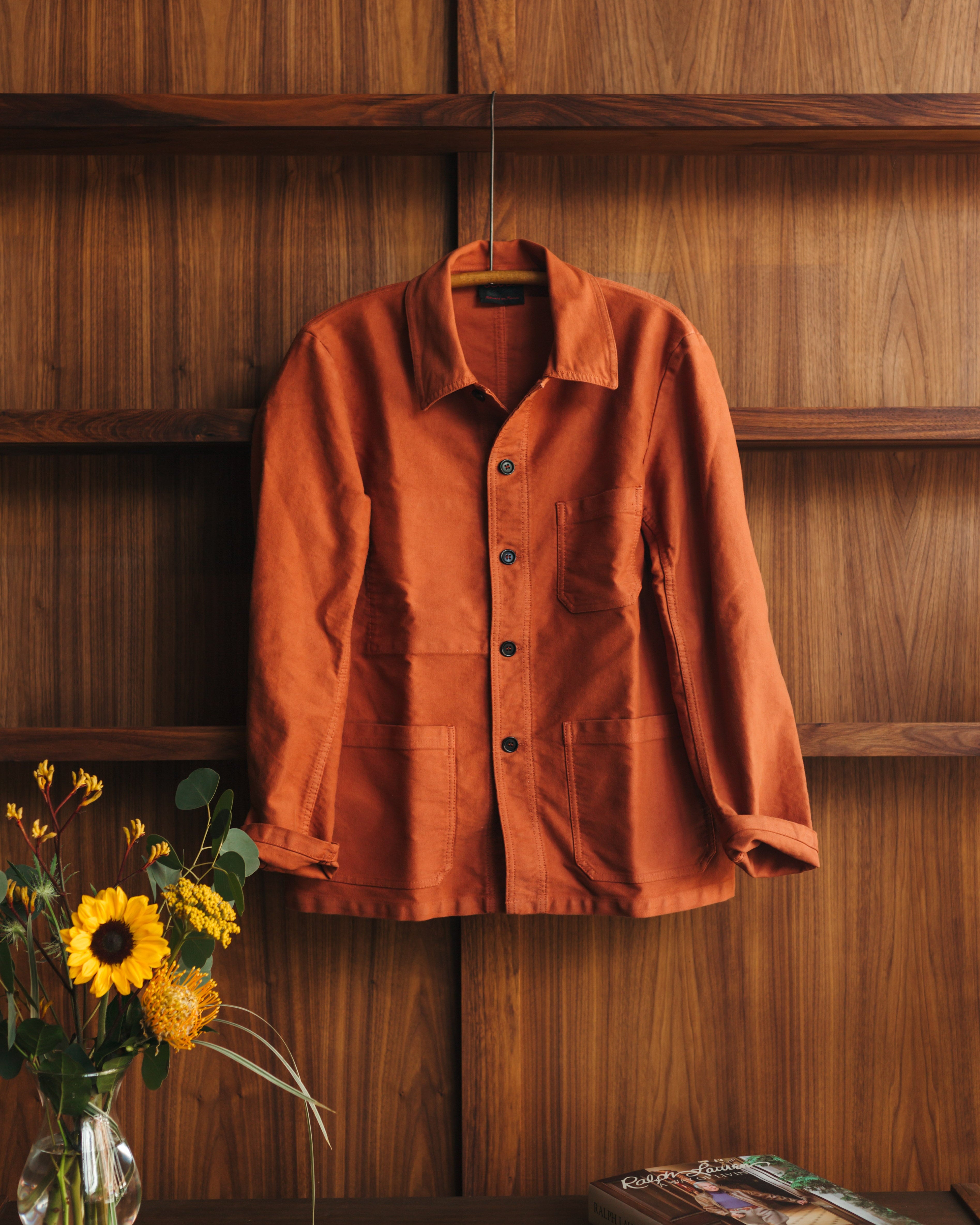Orange Vetra moleskin jacket hanging on a wooden wall with sunflowers and a book below at Fancy Groceries in Winston Salem NC