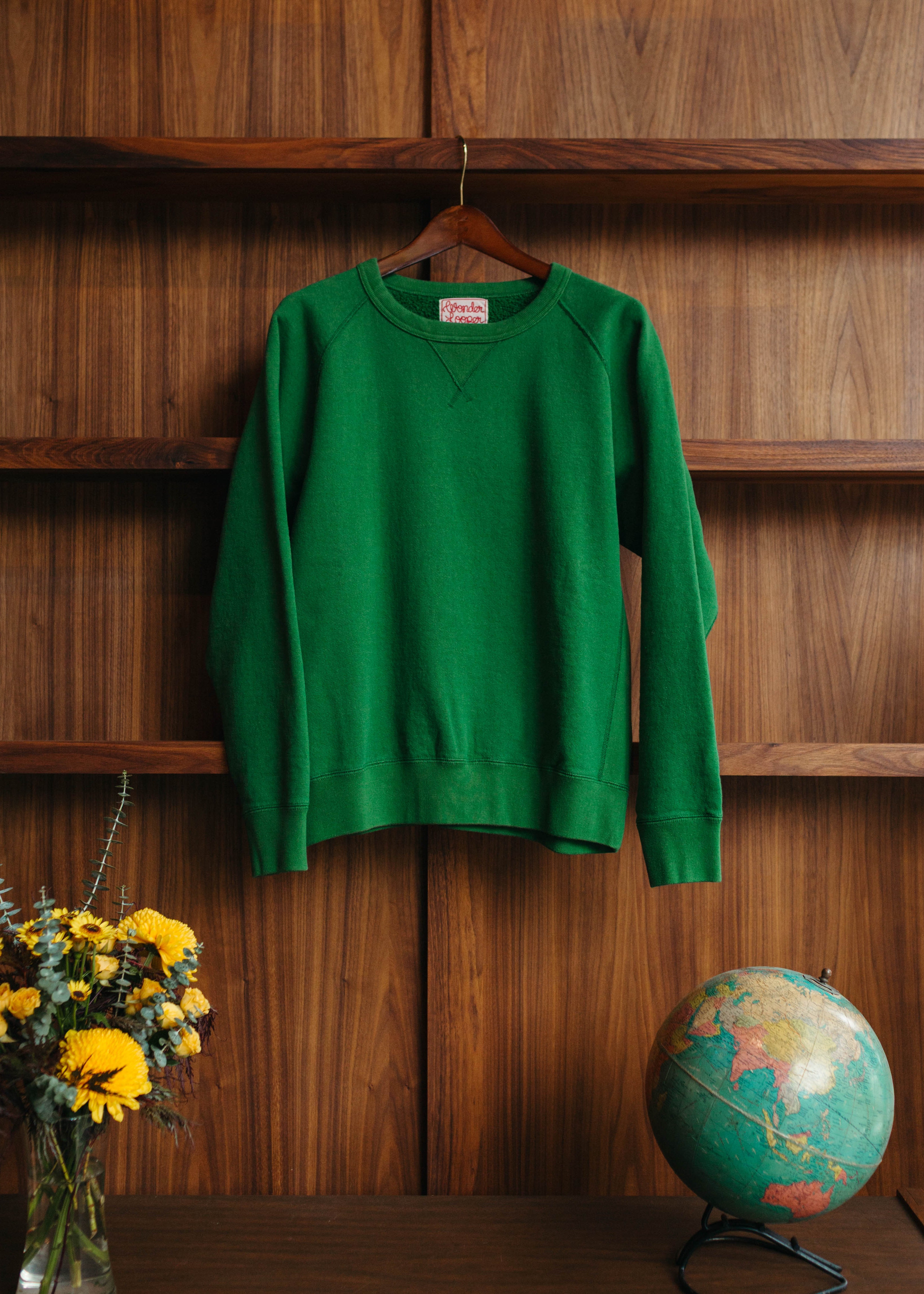 Green sweatshirt hanging on a wooden shelf with a vase of yellow flowers and a globe in the foreground hanging up at Fancy Groceries.